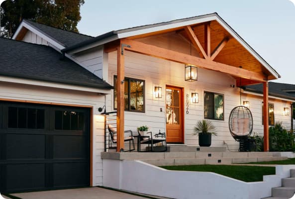 White craftsman home at dusk with a wood-beam porch, orange front door, and warm exterior lighting.