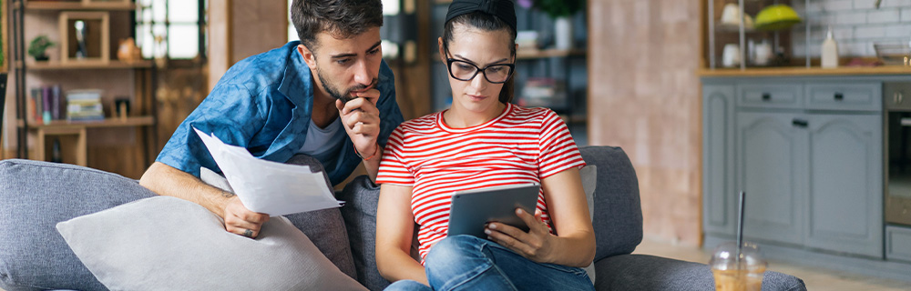 A man and woman sit on a couch with a tablet while reading about ways to save on their electric bill this summer.