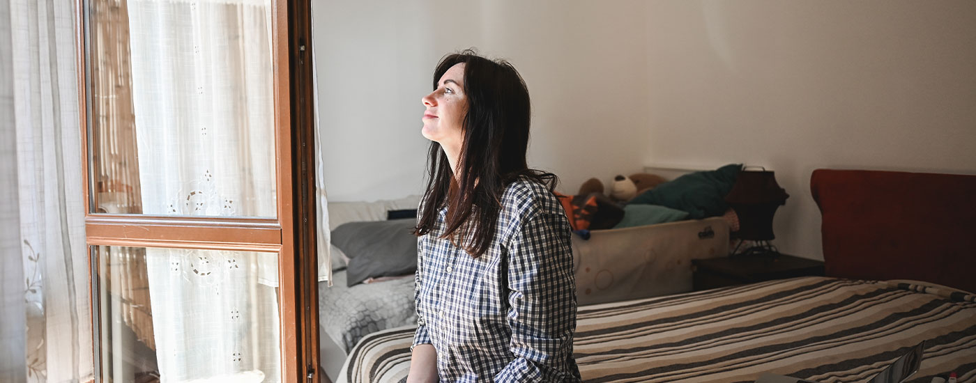 A woman sits inside a small bedroom while gazing out the window of her home.