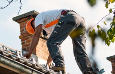 A man in construction gear stands on top of a house to fix the roof.