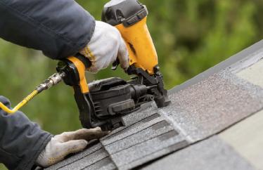 A closeup photo shows someone kneeling on top of a roof while using a tool to lay down new shingles.