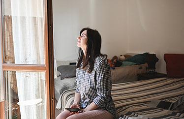 A woman sits inside a small bedroom while gazing out the window of her home.